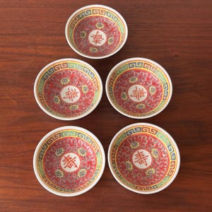 May include: A collection of five small, round ceramic bowls. Each bowl has a red, yellow, and blue decorative pattern and a central Chinese character in red. The bowls are displayed on a dark wooden surface.