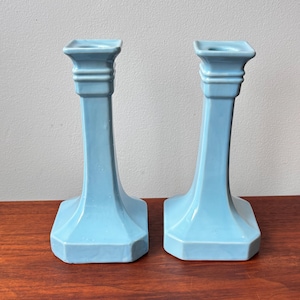May include: A pair of light blue ceramic candle holders. Each has a square top, a long, gently curved stem, and a hexagonal base. The candle holders are displayed on a wooden surface.