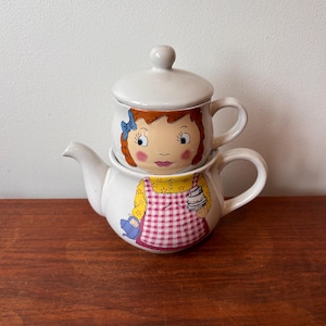 Könnte beinhalten: Ein weißes Keramik-Teeservice, bestehend aus einer Teekanne und einer Tasse. Die Tasse ist auf der Teekanne gestapelt. Beide Teile sind mit einer Cartoon-Illustration eines Mädchens mit roten Haaren, einer blauen Schleife und einer rosa-weiß karierten Schürze verziert.