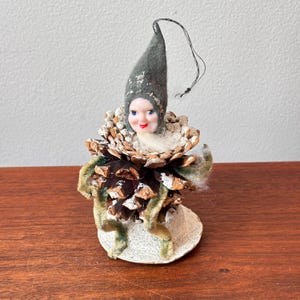May include: A vintage Christmas ornament featuring a whimsical elf figure. The elf has a painted face, a gray felt hat, and a body crafted from a pine cone. Green velvet trim and a white base complete the festive design.