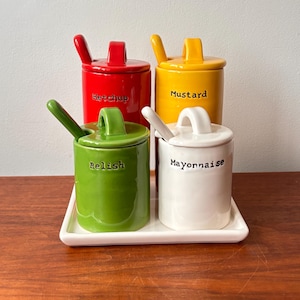 Może przedstawiać: Zestaw czterech ceramicznych słoiczków na przyprawy w kolorze czerwonym (ketchup), żółtym (musztarda), zielonym (relish) i białym (majonez). Każdy słoiczek ma pasującą pokrywkę i łyżeczkę i stoi na białej prostokątnej tacy. Etykiety są w kolorze czarnym.