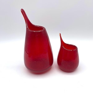 May include: Two red glass vases with a teardrop shape and elongated spouts. The larger vase is approximately 7 inches tall, and the smaller vase is about 4 inches tall. Both vases have a glossy finish and are set against a white background.