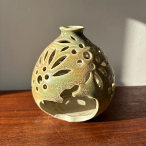 May include: A green ceramic candle holder with a floral design. The holder has a round shape and is made of a textured material.