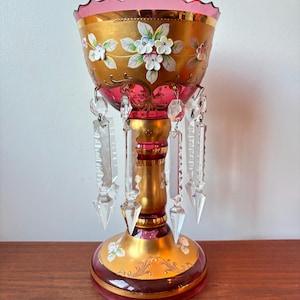 Może przedstawiać: Ozdobny, zabytkowy szklany wazon ze złotą podstawą i misą, ozdobiony różowymi i białymi kwiatowymi wzorami. Wazon posiada wiszące kryształowe zawieszki i falisty brzeg. Wazon to element dekoracyjny.