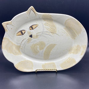 Puede incluir: Una fuente de cerámica con forma de gato. La fuente blanquecina presenta una cara de gato pintada a mano con detalles en marrón y beige. Los ojos del gato son almendrados y los bigotes son líneas finas. La fuente está sobre un soporte metálico dorado.