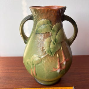 May include: Green ceramic vase with two handles, featuring a floral design with leaves and hanging flowers. The vase has a brown rim and is set on a wooden surface. The design is in relief, adding texture to the vase.