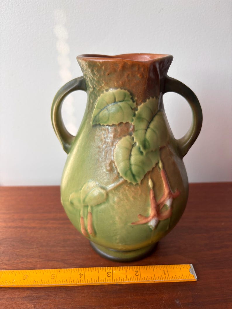 May include: Green ceramic vase with two handles, featuring a floral design with leaves and hanging flowers. The vase has a brown rim and is set on a wooden surface. The design is in relief, adding texture to the vase.