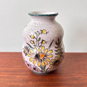 Puede incluir: Un pequeño jarrón de cerámica pintado a mano con acabado craquelado. El jarrón presenta un diseño floral con margaritas amarillas, hojas verdes y detalles morados. El borde y la base están ribeteados en verde. Se muestra sobre una superficie de madera.