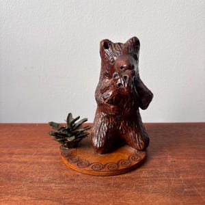 May include: A hand-carved wooden bear figurine in a standing pose, with a detailed texture and rich brown color. The bear is mounted on a decorative wooden base with a carved border. A small pine cone rests beside the bear.