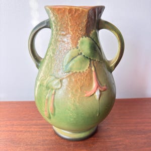 May include: A green and brown ceramic vase with two handles. The vase features a textured surface and a floral design with green leaves and pink flowers. The vase is approximately 20 cm tall.