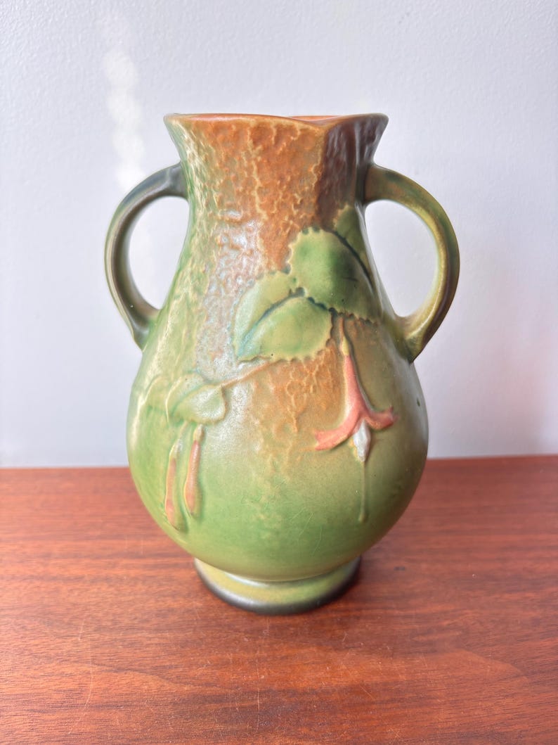May include: A green and brown ceramic vase with two handles. The vase features a textured surface and a floral design with green leaves and pink flowers. The vase is approximately 20 cm tall.