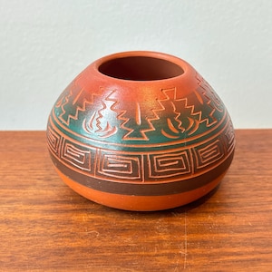 May include: A small, round, terracotta-coloured ceramic pot with a wide opening. The pot features a dark green and brown geometric design around the middle and top, with a lighter orange colour on the upper portion. The design includes geometric shapes and stylized flame-like patterns.