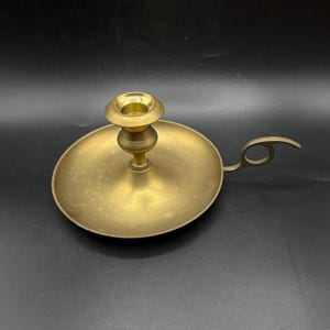 Può includere: Un candelabro in ottone con base rotonda e manico. Il candelabro ha un design semplice e una finitura usurata.