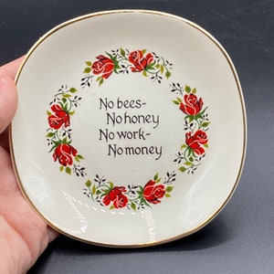 May include: A white ceramic trinket dish with a gold rim and a red rose border. The dish has the text "No bees- No honey No work- No money" in black script.