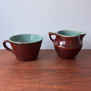 May include: A brown ceramic teacup and creamer set. The cup has a handle and a teal interior. The creamer has a spout and a teal interior. Both pieces are on a wooden surface.
