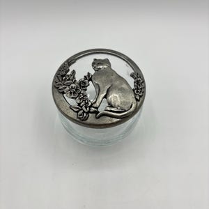 May include: A glass jar with a silver lid featuring a cat sitting in a circle of flowers. The lid is detailed with a raised design of the cat and flowers.