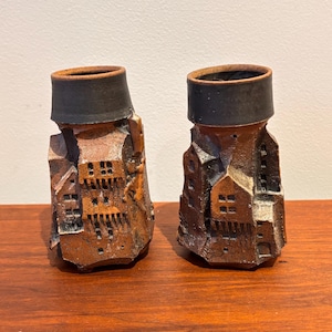 May include: Two brown ceramic vases with a textured, architectural design. Each vase features a dark rim and is sculpted to resemble a cluster of buildings. The vases are displayed on a wooden surface.