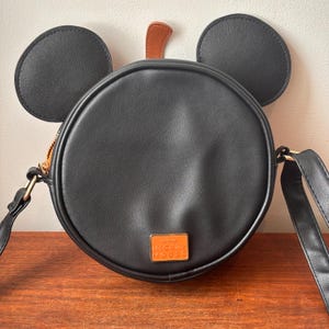 May include: Black round crossbody bag with Mickey Mouse ears and a brown accent. The bag has a gold zipper and a gold rectangular emblem with the words "Disney Mickey Mouse".