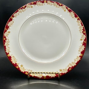 May include: A white ceramic dinner plate with a gold and burgundy rim. The rim features a repeating pattern of gold leaves and vines on a burgundy background.