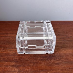 May include: A clear, rectangular glass trinket box with a hinged lid. The box has a geometric design with raised edges and a textured surface. The box is sitting on a wooden surface.