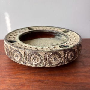 May include: A round, ceramic ashtray with a textured, earthy design. The ashtray features a series of circular patterns around the rim and small holes. The interior has a light beige color, and the exterior is a mix of brown and green.