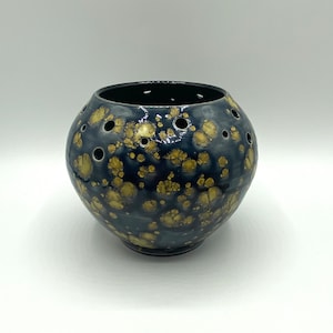 Peut inclure: Un pot en céramique noir avec des taches dorées et des trous partout. Le pot est rond et a une finition lisse et brillante.