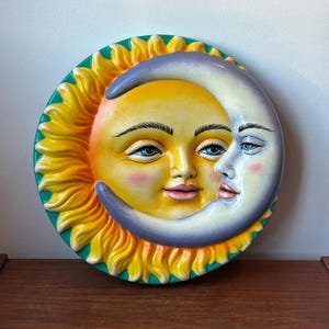 Könnte beinhalten: Eine kreisförmige Wandplatte mit einem gelben Sonnengesicht mit leuchtend blauen Augen und einem violetten Mondsichelgesicht mit blauen Augen, das sich überlagert. Die Sonne hat goldene Strahlen, die bis zu einem blaugrünen Rand reichen, rosa Wangen und rote Lippen.