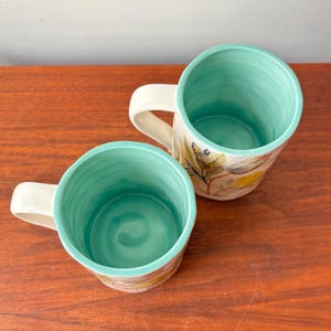 Puede incluir: Dos tazas de cer&aacute;mica con asa color crema e interior turquesa. El exterior presenta un dise&ntilde;o bot&aacute;nico con hojas amarillas y verdes. Las tazas est&aacute;n sobre una superficie de madera.