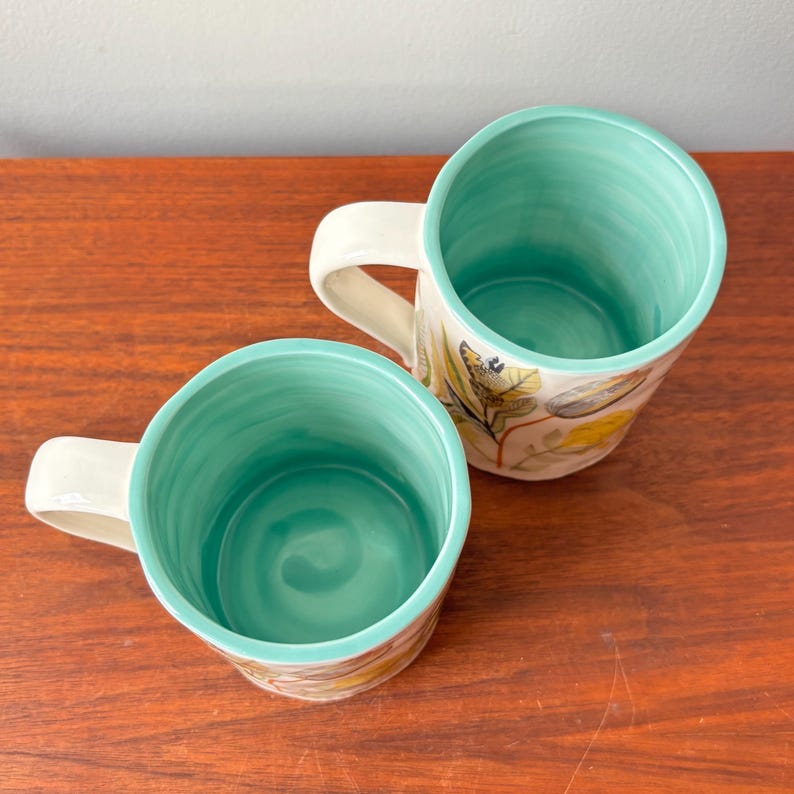 Puede incluir: Dos tazas de cer&aacute;mica con asa color crema e interior turquesa. El exterior presenta un dise&ntilde;o bot&aacute;nico con hojas amarillas y verdes. Las tazas est&aacute;n sobre una superficie de madera.
