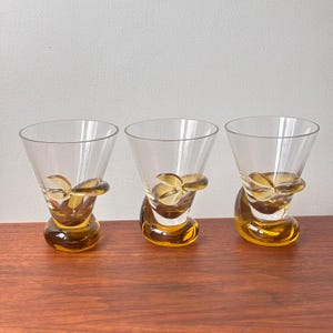 Pode incluir: Três copos de cocktail transparentes com detalhes âmbar. Cada copo tem uma taça aberta e uma base âmbar com um redemoinho decorativo. Os copos estão dispostos sobre uma superfície de madeira.