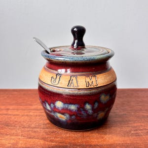 以下が含まれることがあります： 蓋と小さなスプーンが付いた陶器製のジャムジャー。ジャーは深い赤色で、中央に「JAM」と書かれたタン色のバンドが付いています。蓋とハンドルは濃い茶色です。