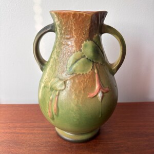 May include: A handcrafted ceramic vase with two handles, featuring a green and brown colour scheme. The vase is adorned with a floral design, showcasing green leaves and pink flower buds. The vase is approximately 20 cm tall.