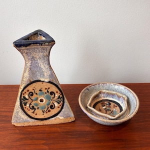 Peut inclure: Ensemble de poterie en céramique fait à la main. Il comprend un vase triangulaire avec un motif floral et deux petits bols. La poterie a un coloris moucheté, violet clair et beige avec des accents bleus et noirs.