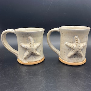 Puede incluir: Dos tazas de cerámica con un esmalte blanco y detalles marrones. Cada taza presenta un diseño de estrella de mar en relieve.