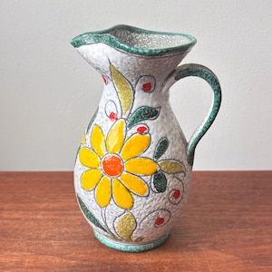 Peut inclure: Pichet en céramique avec une surface blanche texturée, orné d'une grande fleur jaune avec un centre orange. Des accents verts et rouges ornent le motif floral. La poignée et le bord sont verts. Le pichet est posé sur une surface en bois.