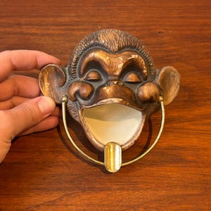 May include: A vintage monkey-shaped ashtray, made from brown and gold ceramic, with a smiling face. It has a gold-coloured metal handle and a white interior. The ashtray is designed with a slot for cigarettes.