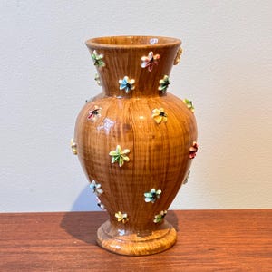 May include: A brown ceramic vase with a glossy finish, decorated with colourful, raised flower accents. The vase has a bulbous body, a narrow neck, and a flared base. The flowers are in shades of red, yellow, blue, and green.