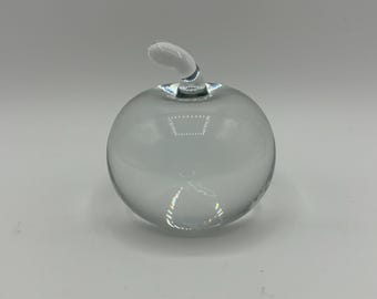 May include: A clear glass apple sculpture with a curved stem. The apple is smooth and round, reflecting light. The sculpture is a decorative item, suitable for display on a shelf or table.