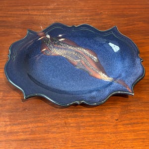 Peut inclure: Plat ovale en céramique émaillée bleu foncé. Le plat présente un motif décoratif avec des accents bruns et crème. Le bord est festonné, ajoutant à son attrait artistique. Idéal pour servir ou décorer. Dimensions: environ 25 cm de long.