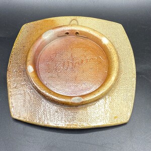 K&ouml;nnte beinhalten: Ein quadratischer Keramikteller mit brauner und beigefarbener Glasur. Der Teller hat eine runde, erhabene Mitte mit brauner Glasur und das Wort "Barbier" in Kursivschrift. Der Teller hat einen Metallb&uuml;gel auf der R&uuml;ckseite.