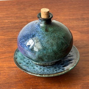 Puede incluir: Una aceitera de cerámica redonda con un esmalte brillante en tonos azul profundo, verde azulado y verde, con un cuello estrecho rematado por un tapón de corcho cilíndrico de color marrón claro. La aceitera descansa sobre un plato de cerámica a juego.