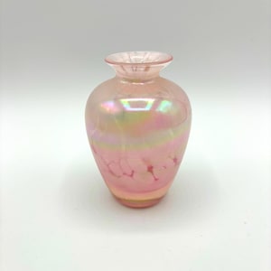 Peut inclure: Un petit vase en verre rose nacré, de forme arrondie et au col étroit. Le vase présente un léger reflet irisé, avec des touches de jaune et de vert. La base est d'une nuance de rose plus foncée, et la surface présente un motif délicat et craquelé.