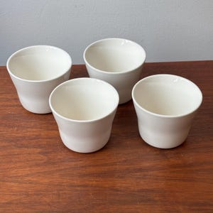 May include: A set of four off-white ceramic cups with a smooth, rounded silhouette and a wide, open top. Each cup features a simple, handleless design with a slightly flared rim and a solid, uniform color throughout the exterior and interior.
