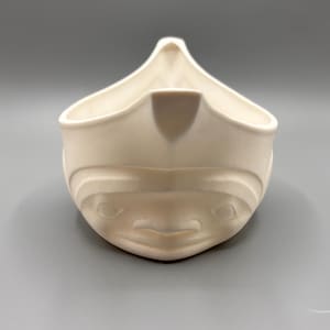 Pode incluir: Uma tigela de cerâmica branca em forma de canoa com um rosto esculpido na frente.
