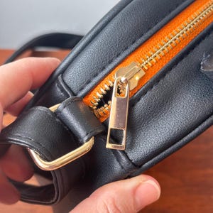 May include: Close-up of a black crossbody bag with a gold zipper and hardware. The interior lining is bright orange. The bag is made of a smooth, leather-like material.