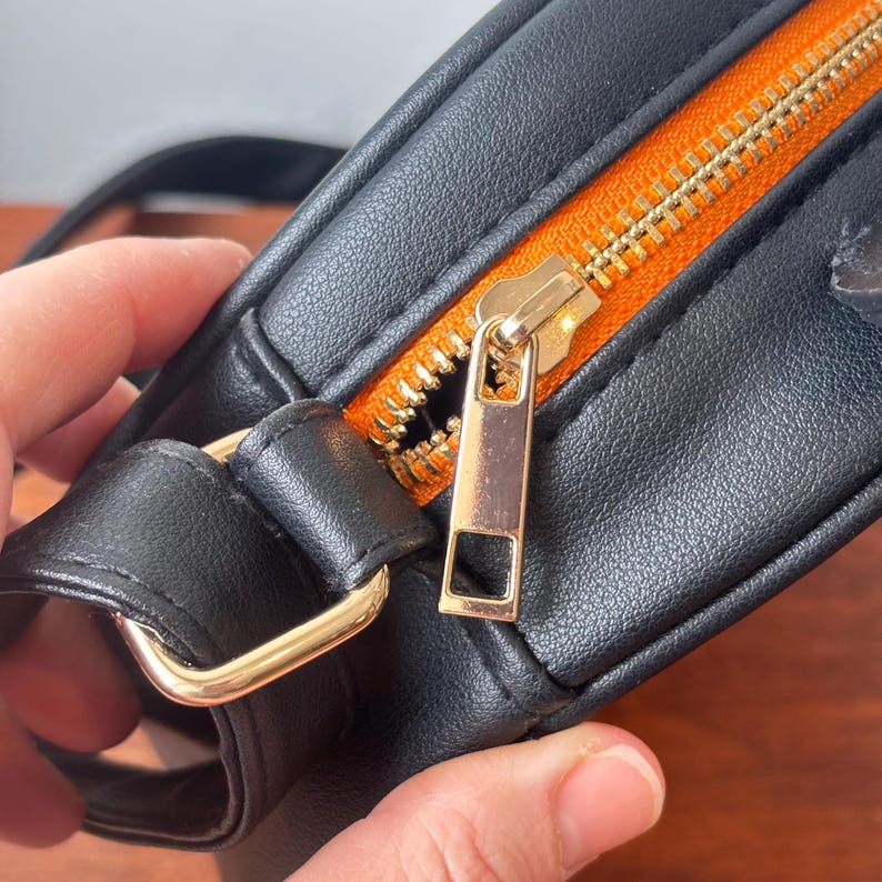 May include: Close-up of a black crossbody bag with a gold zipper and hardware. The interior lining is bright orange. The bag is made of a smooth, leather-like material.