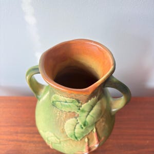 May include: A green ceramic vase with two handles and a wide opening. The vase features embossed leaf designs and a brown interior. The exterior is a muted green with brown accents, creating a vintage aesthetic.