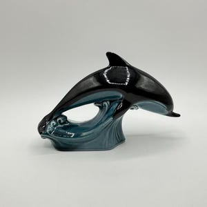 Può includere: Statuetta di orca in ceramica nera e verde acqua. L'orca è raffigurata mentre salta sopra un'onda verde acqua. La statuetta è un oggetto decorativo, probabilmente per una mensola o un'esposizione.