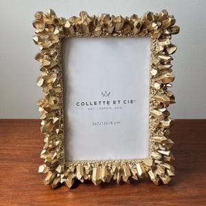 May include: A rectangular picture frame with a metallic gold finish and a textured, crystalline border. The white insert features the black text COLLETTE ET CIE EST. DEPUIS 2015 and 5x7/13x18 cm. The frame has a jagged, rock-like appearance.