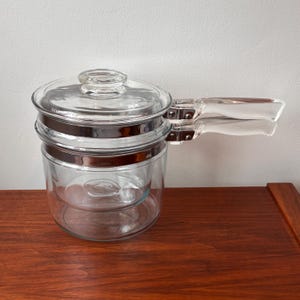 Puede incluir: Una olla de doble caldera de vidrio transparente con tapa y asa transparente. La olla tiene una banda plateada alrededor del centro y un asa de vidrio transparente. La olla de doble caldera está sobre una superficie de madera.